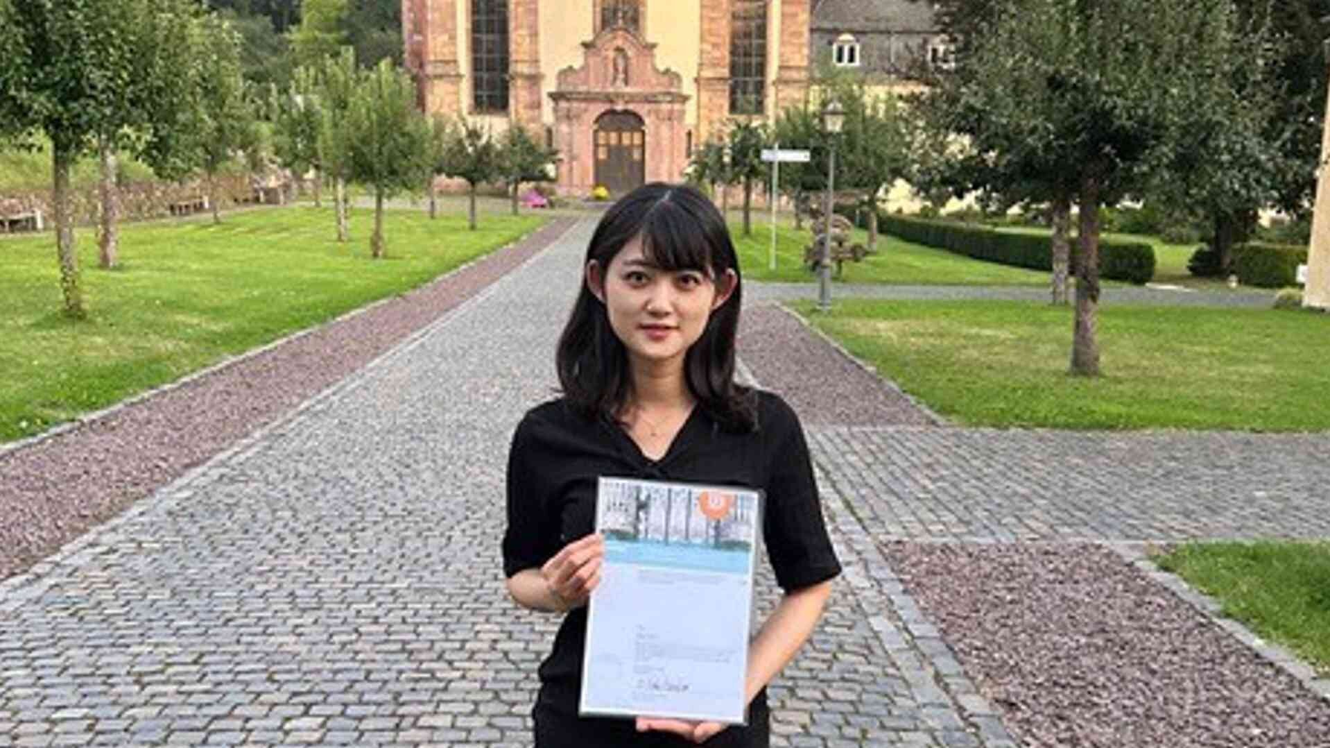 Risa Toho steht mit Urkunde in der Hand vor einer Kirche