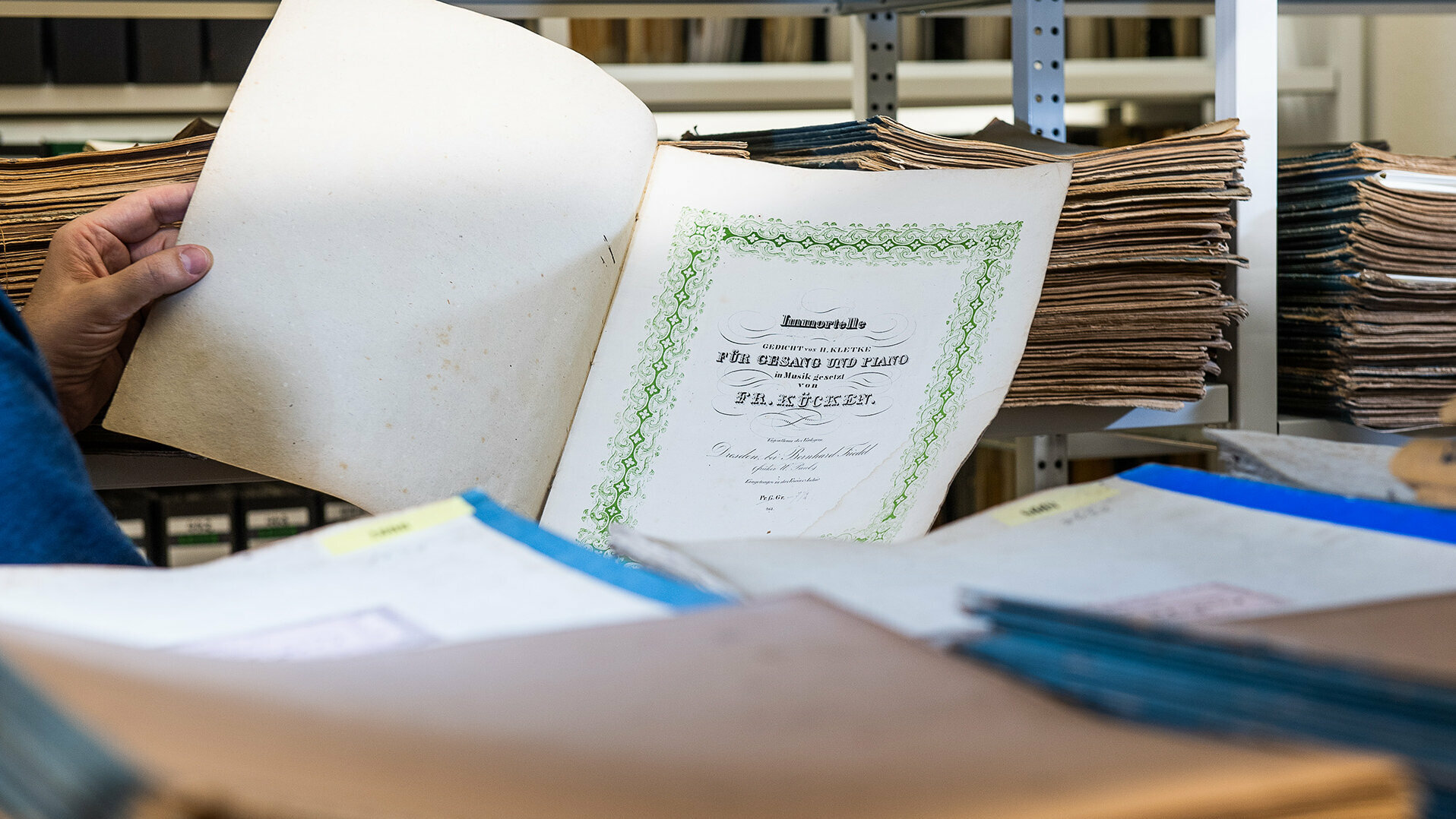 Aufgeschlagene Noten umgeben von Papierstapeln in einem Archiv 